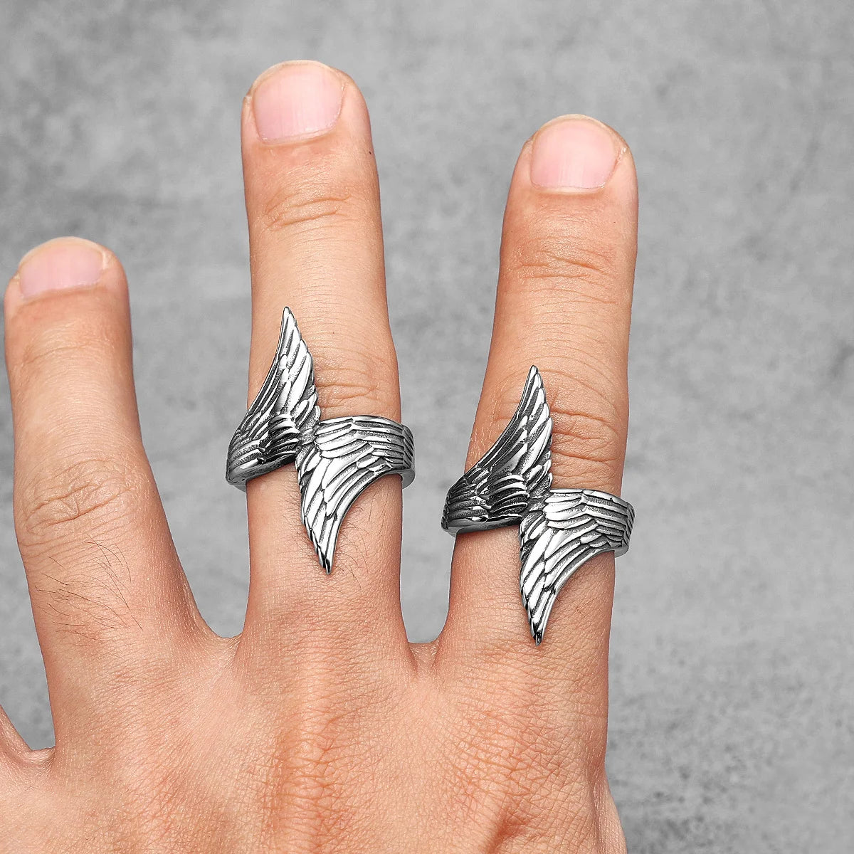 Silver wing-shaped rings on a person's fingers against a grey background