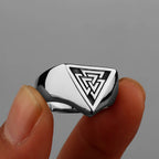 Silver ring with a valknut design held between fingers against a grey background