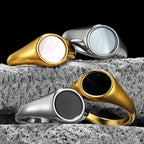 Four simple pearl shell rings in gold, silver, and black on a textured stone surface with a black background.