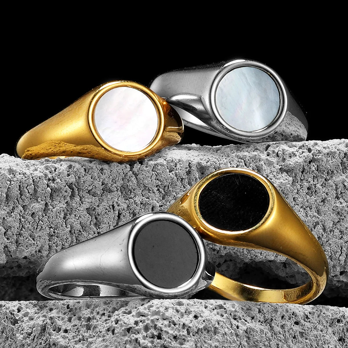Four simple pearl shell rings in gold, silver, and black on a textured stone surface with a black background.
