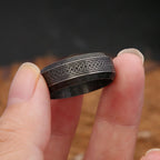 Dark Grey ring with intricate Nordic design held between fingers against a blurred background