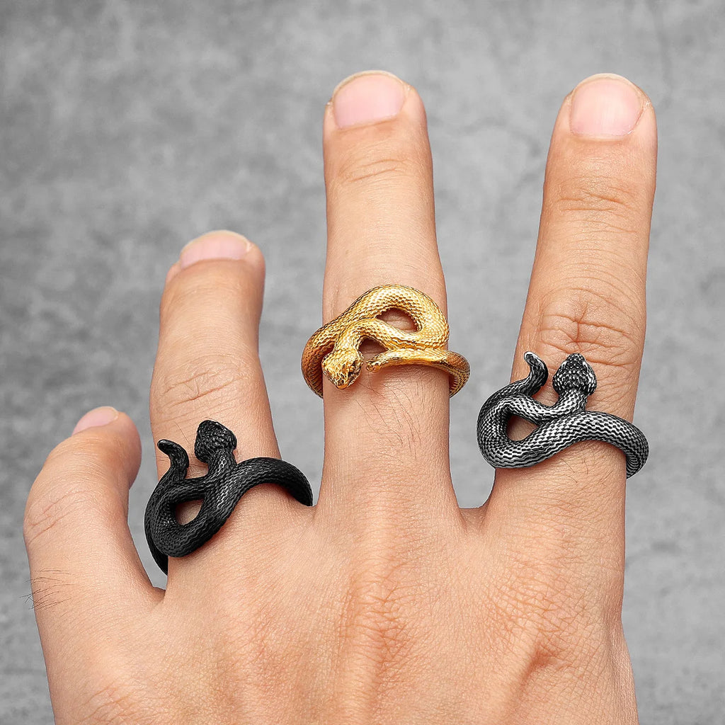Hand wearing three snake-shaped rings in black, gold, and silver on a grey background
