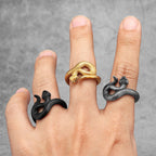 Hand wearing three snake-shaped rings in black, gold, and silver on a grey background