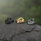 three snake and viper detailed shaped rings of gold, silver and black sitting on a dark grey rock with blurred green background