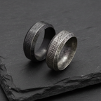 Two silver rings with Celtic knot and Norse runic designs on a dark stone charcoal surface