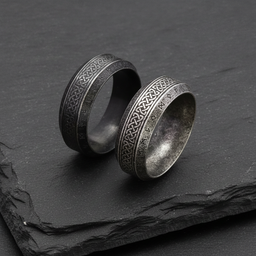 Two silver rings with Celtic knot and Norse runic designs on a dark stone charcoal surface