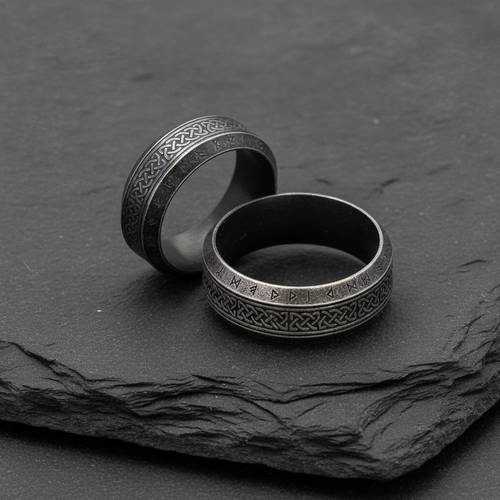 Two stainless steel rings with intricate Norse designs on a dark charcoal stone surface