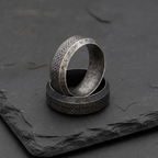 Two silver stainless steel rings with intricate Norse patterns on a dark stone surface