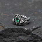 Silver ring with a green artificial gemstone and twin serpent design on a dark basalt stone surface